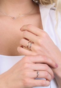 Zilveren ringen met gepolijste oppervlakken; de een is een gladde band, de andere heeft een zigzagontwerp. Handen zijn gepositioneerd bij een zilveren hartketting.