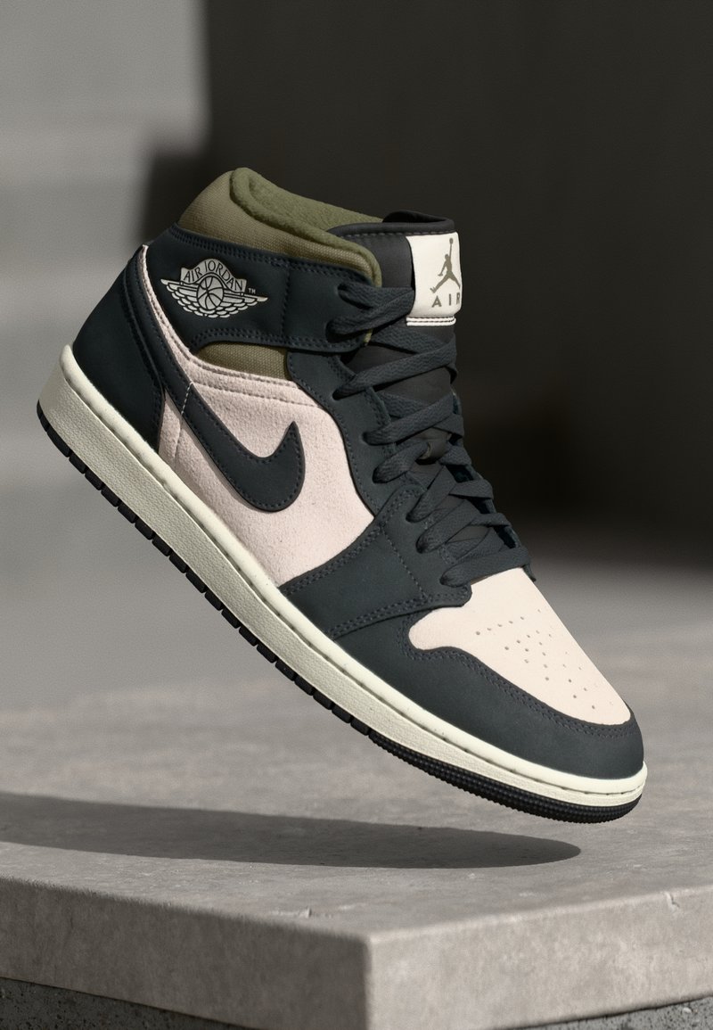 Basket Air Jordan 1 montante mi-haute en noir, blanc et vert olive suspendue au-dessus d'une surface en béton.