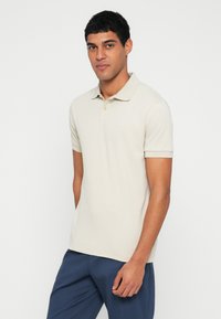 Polo bege com textura suave, mangas curtas e gola clássica. Apresenta um fecho com um único botão e um ajuste casual.
