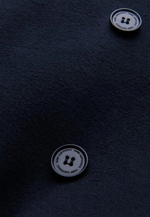 Marineblaue Stoff mit weicher Textur, der mit zwei runden, schwarzen Knöpfen versehen ist, von denen jeder ein Logo und vier kleine Löcher aufweist, die auffällig platziert sind.