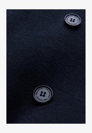 Tessuto blu navy con una texture morbida presenta due bottoni rotondi neri, ciascuno con un logo e quattro piccole aperture, posizionati in modo evidente.