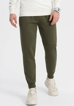 Olivgrüne Sweatpants mit gerippter Textur, schmalem Schnitt und einem horizontalen Akzentstreifen am Knie. Kombiniert mit hellen Turnschuhen.