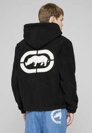 Mladá osoba nosí čiernu fleecovú mikinu s kapucňou a bielym logom nosorožca a svetlomodré rifle s nášivkou nosorožca na zadnom vrecku.