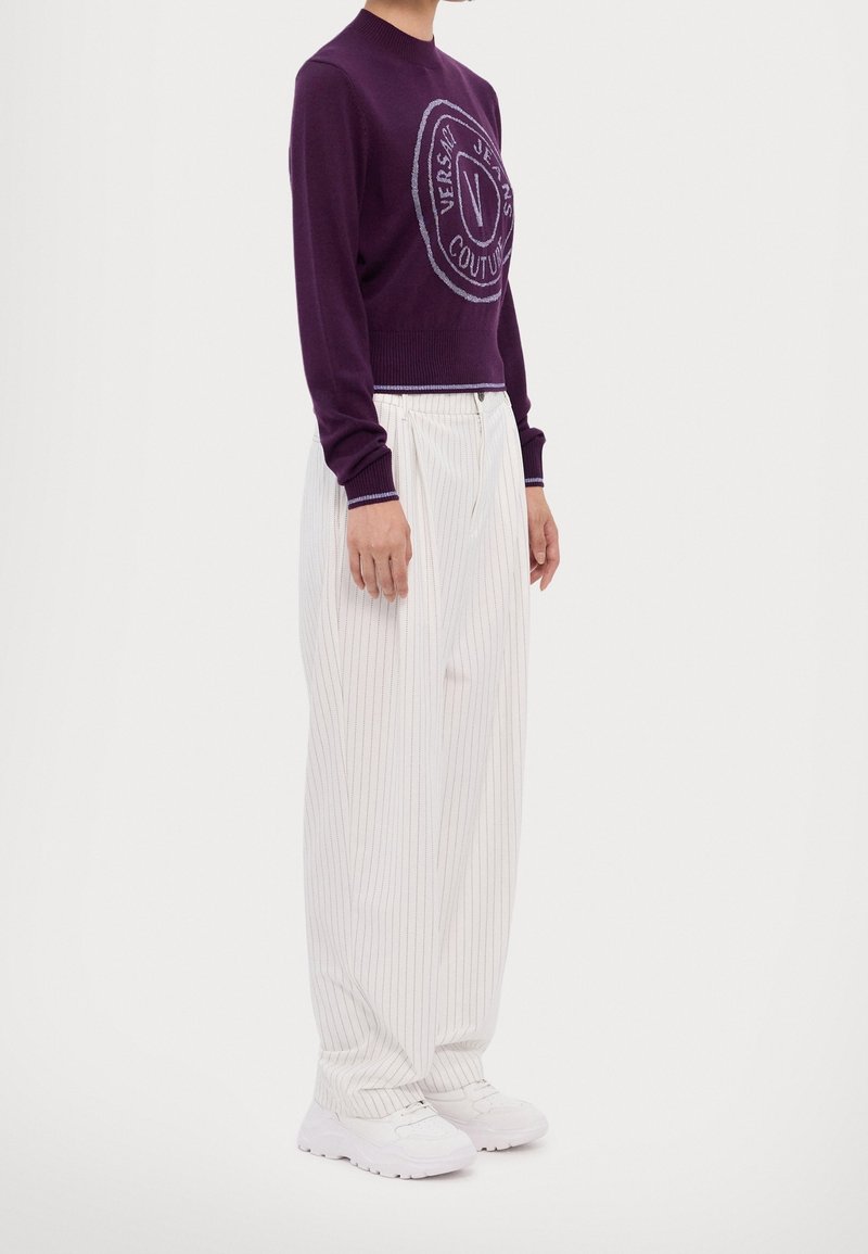 Personne portant un pull violet Versace Jeans Couture, un pantalon large à fines rayures blanches, et des baskets blanches, debout devant un fond uni.