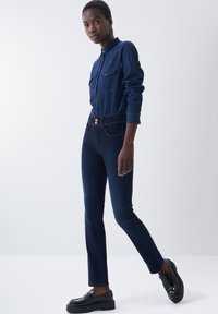 Camisa de mezclilla azul oscuro con dos bolsillos en el pecho, combinada con unos vaqueros ajustados azul oscuro y zapatos de charol negros, que cuenta con un cierre de dos botones.