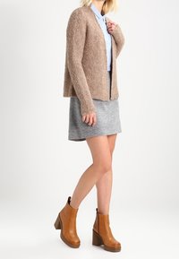 Cardigan en maille beige sur une chemise bleu clair, associé à une jupe grise et des bottines à talons bruns. Matériaux texturés et design ajusté.