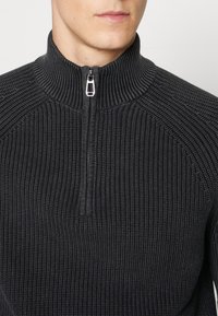 Czarna bluza z suwakiem i wysokim kołnierzem, o prążkowanej fakturze i dopasowanym kroju. Suwak z detalem srebrnego okucia.