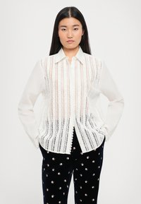 Bílá krajková blůza s vertikálním pruhovaným detailem, zapínáním na knoflíky a dlouhými rukávy, v kombinaci s černými sametovými kalhotami se květinovými vzory.