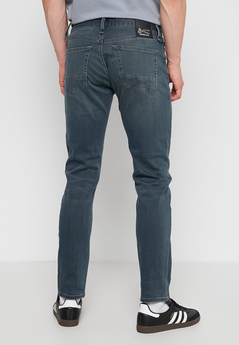 Donkerblauwe denim jeans met een slim fit, voorzien van twee achterzakken en een lage taille, gecombineerd met zwarte sneakers en witte accenten.