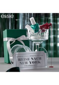 Bol en verre cristal contenant trois flacons de vernis à ongles (rouge et teal) et un billet promotionnel en argent sur une surface grise texturée, à côté d'une boîte cadeau verte.