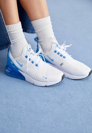 Baskets Nike Air Max 270 blanches et bleues, portées avec des chaussettes blanches côtelées et un pantalon en jean court sur un tapis bleu clair.