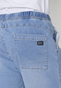 Heledsinised denimpüksid elastse vöökohtaga, millel on tagatasku ja silt. Kanga pind on sile ja tekstuuriga.