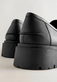 Zapatos de cuero negro con un acabado texturizado, suelas negras gruesas y un diseño elegante. La vista se enfoca en la parte trasera y los lados.
