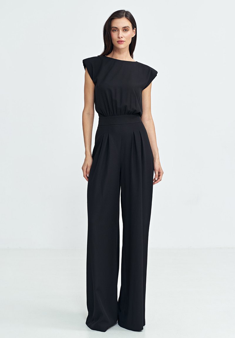 Czarny kombinezon uszyty z gładkiego materiału, z szerokimi poduszkami na ramionach, wcięciem w talii oraz szerokimi spodniami z plisami.