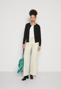 Zwarte cardigan met gouden knopen over een witte top, gecombineerd met crèmekleurige, hoge taille brede broek en zwarte enkelboots; houdt een teal sjaal vast.