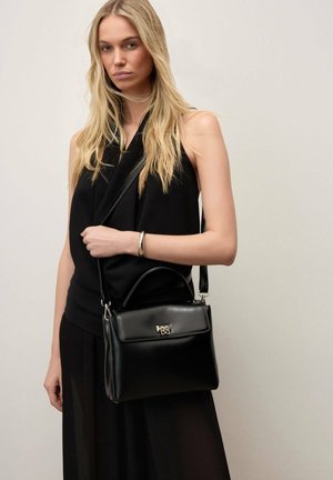 Szőke nő fekete ujjatlan felsőt és szoknyát visel, kezében fekete bőr kézitáskával, amely vállpánttal és ezüst kapoccsal rendelkezik.