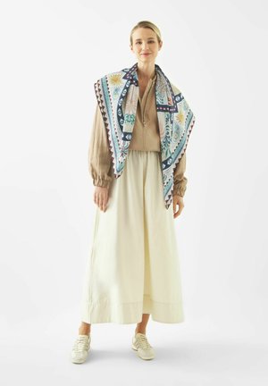 Femme portant un chemisier beige, une jupe large crème, un foulard coloré à motifs drapé sur les épaules, et des baskets blanches, debout devant un fond blanc.