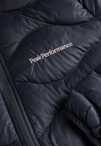 Navy blue puffer jacket featuring stitched design, zip closure, and "Peak Performance" logo in silver thread on the chest. Soft, lightweight material.