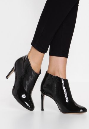 Bottines en cuir verni noir à talons aiguilles hauts, portées avec un pantalon noir ajusté sur un fond blanc.