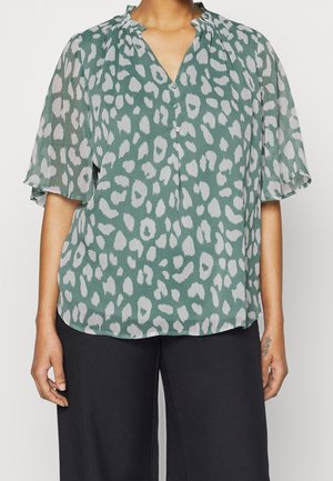 Femme portant un chemisier vert avec des taches grises irrégulières, des manches courtes volantées et un pantalon noir à jambes larges, debout les bras détendus.