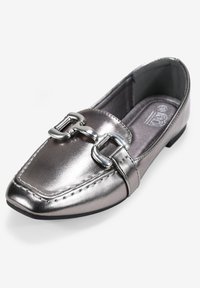 Mocasin metálico plateado con un diseño elegante, que presenta una hebilla decorativa plateada y detalles de costura en la punta redondeada.