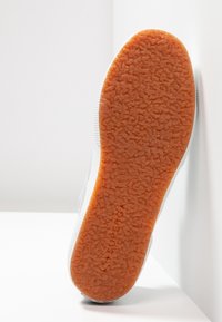 Baskets blanches avec une tige en tissu texturé et une semelle en caoutchouc orange. La semelle présente un motif antidérapant. Le nom de la marque est visible sur la semelle.