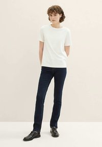 Bela majica s kratkimi rokavi in okroglim izrezom, združena z temno modrimi jeans hlačami z ravnimi hlačnicami. Model nosi črne čevlje in stoji pred svetlim ozadjem.