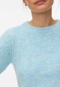 Približen posnetek osebe, ki nosi svetlomodro puloversko majico iz mehke pletene tkanine in srebrno obročno uhan, na belem ozadju.