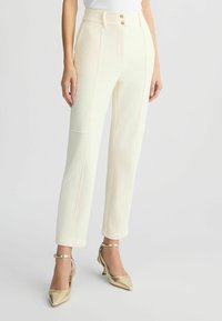 Pantaloni tailored color crema con due tasche frontali, cuciture a contrasto e chiusura con doppio bottone. Abbinati a tacchi a punta dorati.