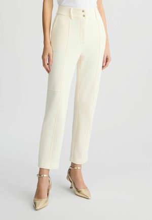 Cream-colored tailored pants with two front pockets, contrasting seams, and dual button closure. Paired with gold pointed-toe heels.