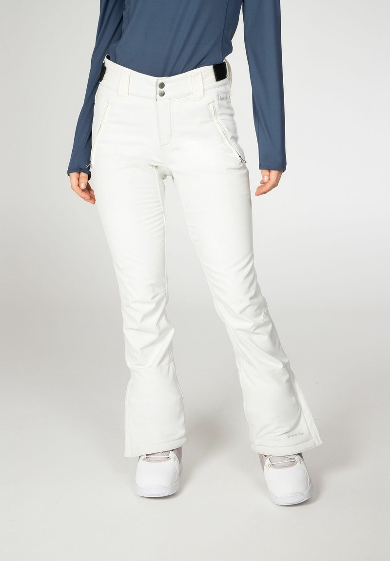 Femme portant un pantalon de neige blanc avec poches zippées, un haut à manches longues bleu marine et des bottes d'hiver blanches, debout sur un fond clair.