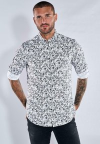 Weißes Hemd mit schwarzem Paisleymuster, mit Knopfkragen und hochgerollten Ärmeln. Modell trägt schwarze Hose, Hintergrund ist neutral grau.