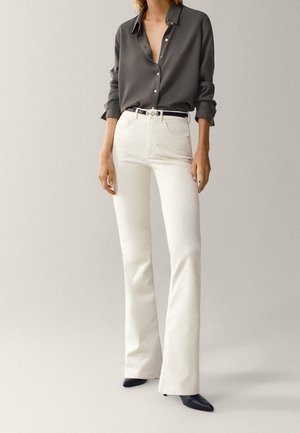 Frau trägt ein dunkelgraues, langärmliges Hemd mit Knöpfen, das in hoch taillierte weiße Schlagjeans gesteckt ist, dazu schwarze spitze Schuhe.