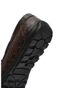 Brauner Lederschuh mit Krokodilstruktur, aus schräger Sohlenansicht gezeigt, mit dickem schwarzem Profil und kreisförmigen sowie rechteckigen Mustern.