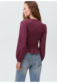 Burgunderfarbenes Oberteil mit gerafftem Oberteil, versammelten Puffärmeln und Rüschensaum; kombiniert mit hellblauen Jeans.