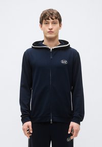Sudadera con capucha azul marino con cremallera y capucha forrada en gris contrastante, con un parche de logo en el pecho y puños acanalados. Tejido suave, ajuste casual.