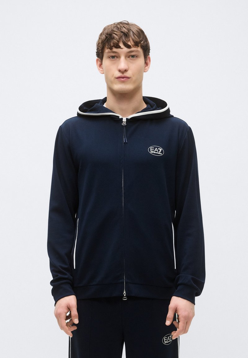 Sudadera con capucha azul marino con cremallera y capucha forrada en gris contrastante, con un parche de logo en el pecho y puños acanalados. Tejido suave, ajuste casual.