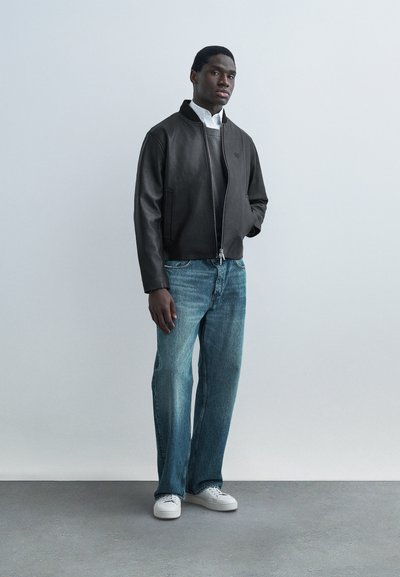 Homme debout les mains dans les poches, portant une veste en cuir noire, une chemise blanche, un jean bleu et des baskets blanches, sur un fond clair uni.