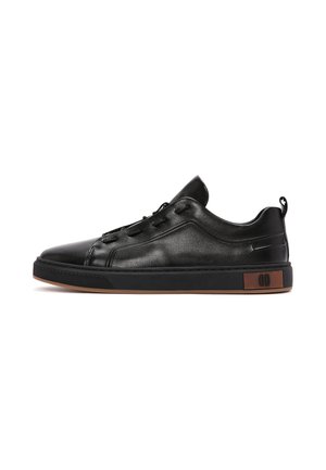 Zwarte lage leren sneaker met zwarte zool, bruine accenten op de hiel, zijstiksels en een treklus aan de achterkant.