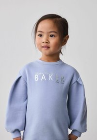Ragazzina dai capelli scuri raccolti, che indossa un maglione azzurro chiaro con la scritta "BAKER", che guarda leggermente di lato su uno sfondo semplice.