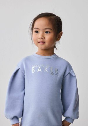 Jeune fille aux cheveux bruns attachés, portant un pull bleu clair avec le texte "BAKER", regardant légèrement sur le côté devant un fond uni.