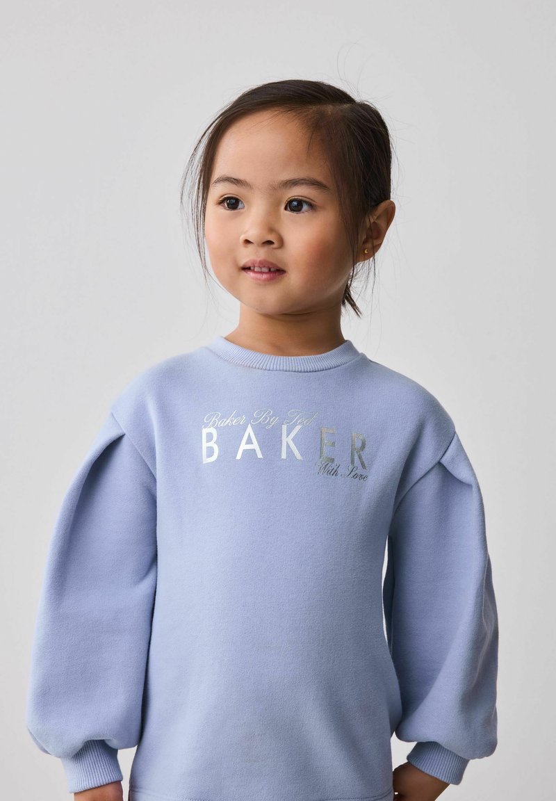 Ragazzina dai capelli scuri raccolti, che indossa un maglione azzurro chiaro con la scritta "BAKER", che guarda leggermente di lato su uno sfondo semplice.