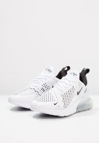 Pár bílých tenisek Nike Air Max 270 s černými detaily a průhlednou vzduchovou podrážkou, zobrazených pod mírným úhlem na bílé ploše.