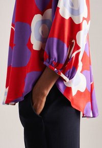 Bunte Bluse mit roten, lila und weißen Blumenmustern. Weicher Stoff, lockere Passform, abgerundeter Saum und Knopfdetail an der Manschette.