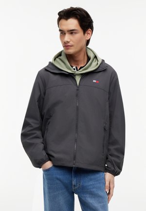 Ung mand iført en sort lynlåstrøje over en olivengrøn hættetrøje, med hænderne i lommerne, kiggende let til siden.