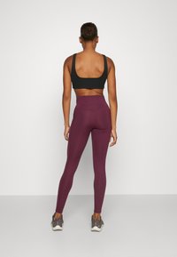 Fekete sportmelltartó és magas derekú bordó leggings, testhezálló kialakítás, sima textúra, szürke sportcipővel párosítva. Tiszta háttér.