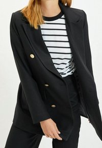 Svart dubbelknäppt blazer med guldknappar, med klassisk slag. Bärs över en randig skjorta, slät tyg, skräddarsydd passform.