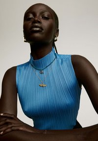 Top sin mangas azul con pliegues y cuello alto. La modelo lleva collares de oro en capas y con cuentas, que incluyen un colgante con alas y pendientes de aro dorados.