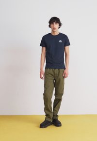 Navy bomull T-skjorte med en liten hvit logo, olivegrønne bukser og svarte sko. Enkel design, tettsittende fasong, glatt tekstur.