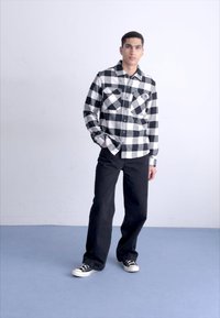 Camisa de franela a cuadros en blanco y negro con dos bolsillos en el pecho, combinada con pantalones anchos negros y zapatillas negras sobre un suelo azul.
